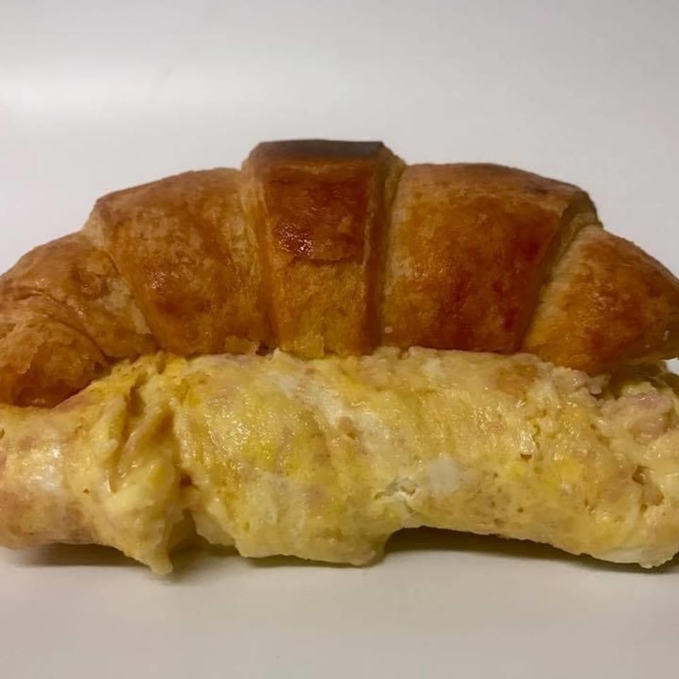 Golden-brown croissant sandwich filled with fluffy scrambled eggs and ham on a white background.