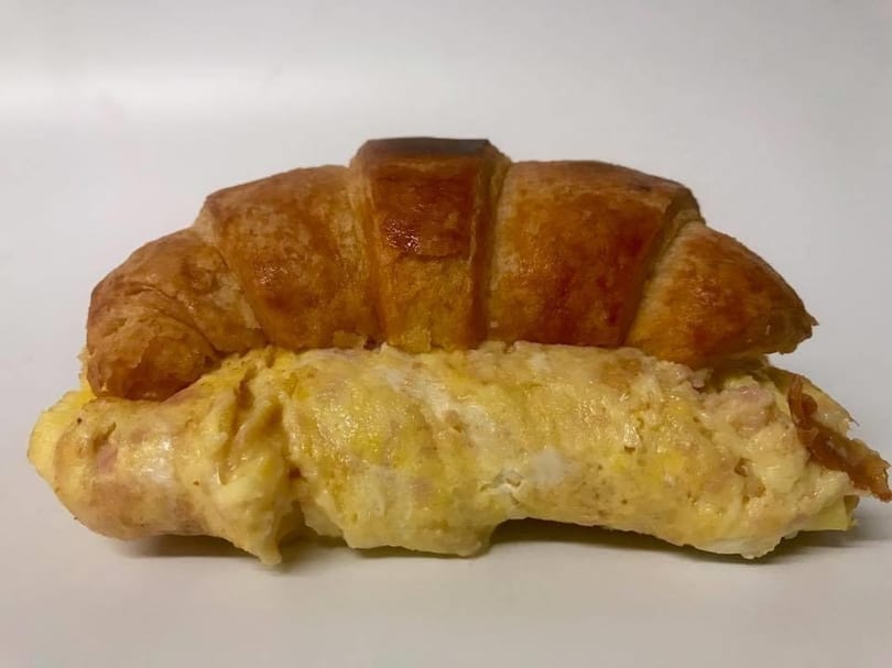Golden-brown croissant sandwich filled with fluffy scrambled eggs and ham on a white background.