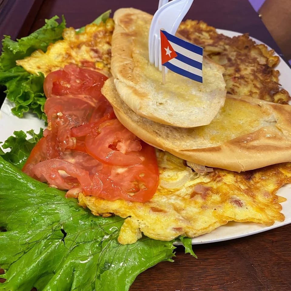Cuban breakfast plate with omelet, toasted bread, sliced tomatoes, lettuce, and a Cuban flag pick.