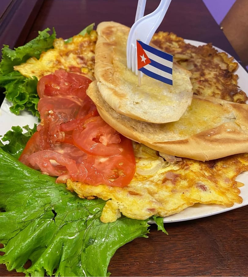 Cuban breakfast plate with omelet, toasted bread, sliced tomatoes, lettuce, and a Cuban flag pick.
