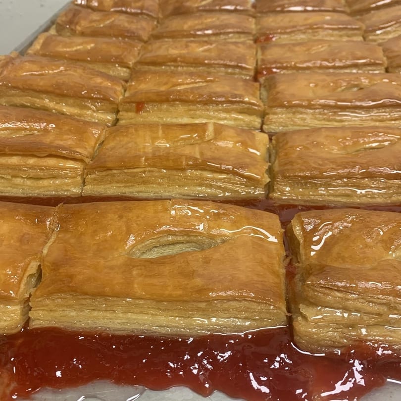 Rows of golden-brown pastries with flaky layers, glistening with syrup, sitting in a pool of red jam.