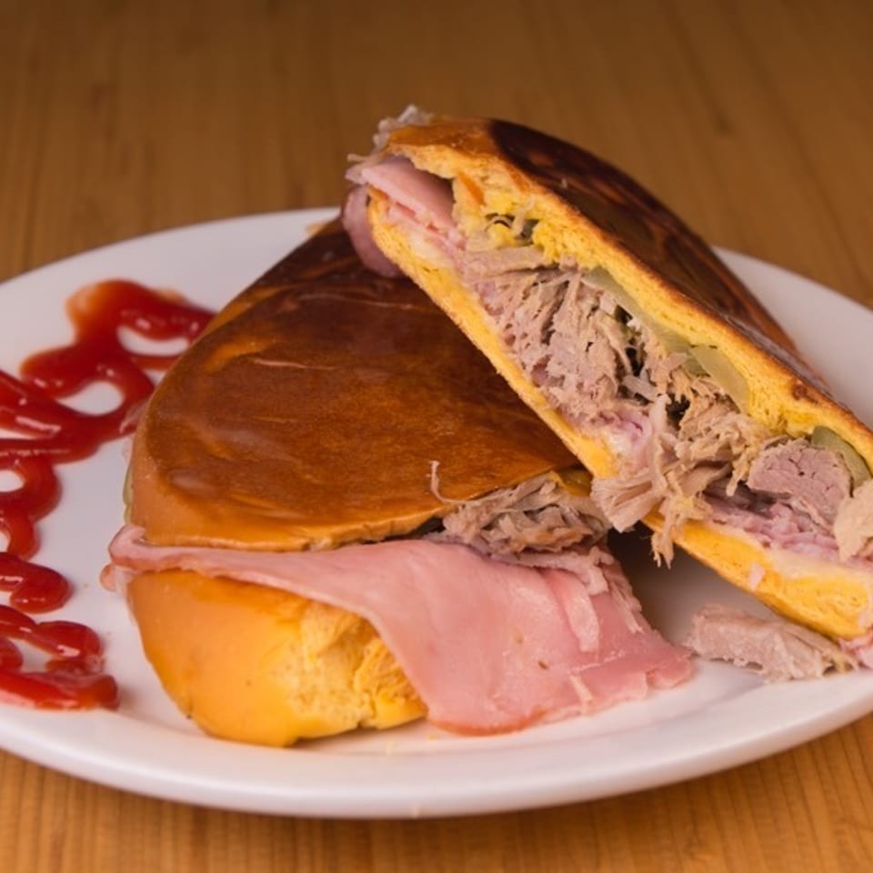 A Cuban sandwich, cut in half, revealing ham, roasted pork, cheese, and pickles, with ketchup on a white plate.