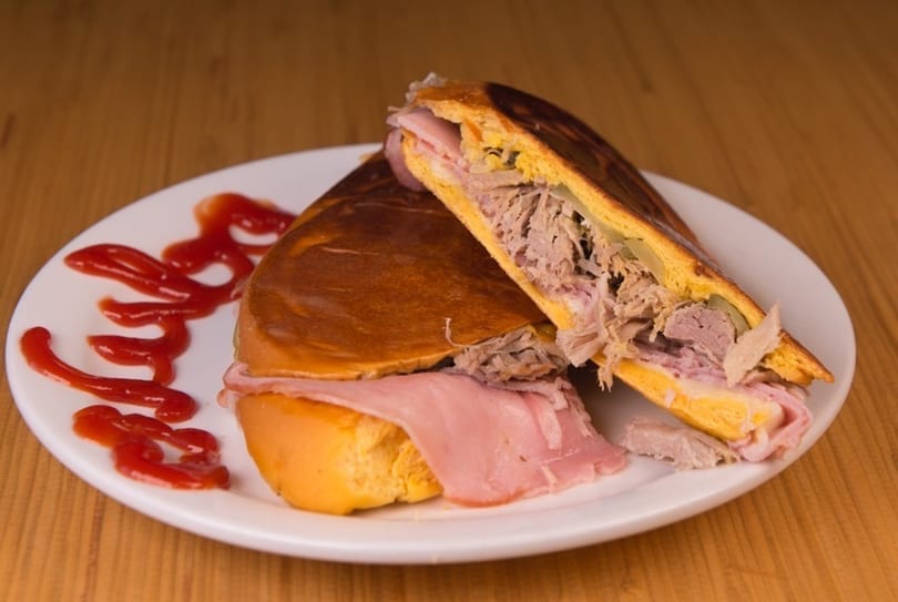 A Cuban sandwich, cut in half, revealing ham, roasted pork, cheese, and pickles, with ketchup on a white plate.