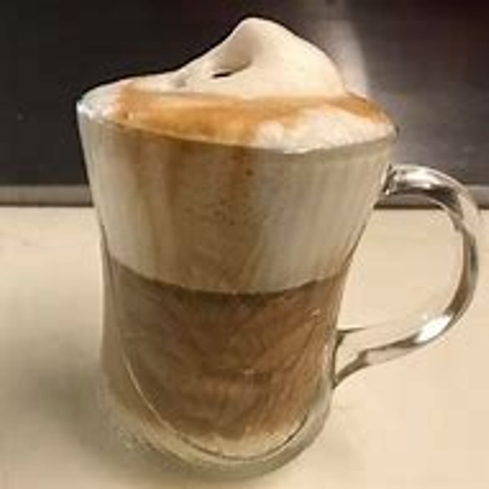 A clear glass mug filled with a layered latte, topped with a generous swirl of frothy milk.