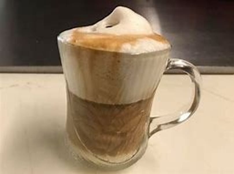 A clear glass mug filled with a layered latte, topped with a generous swirl of frothy milk.