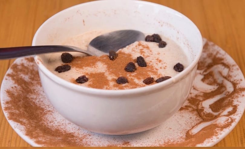 A white bowl of creamy oatmeal topped with cinnamon and raisins, with a spoon resting inside.