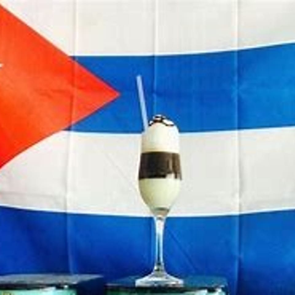 Layered white and brown drink with a straw in a tall glass, against a Cuban flag.