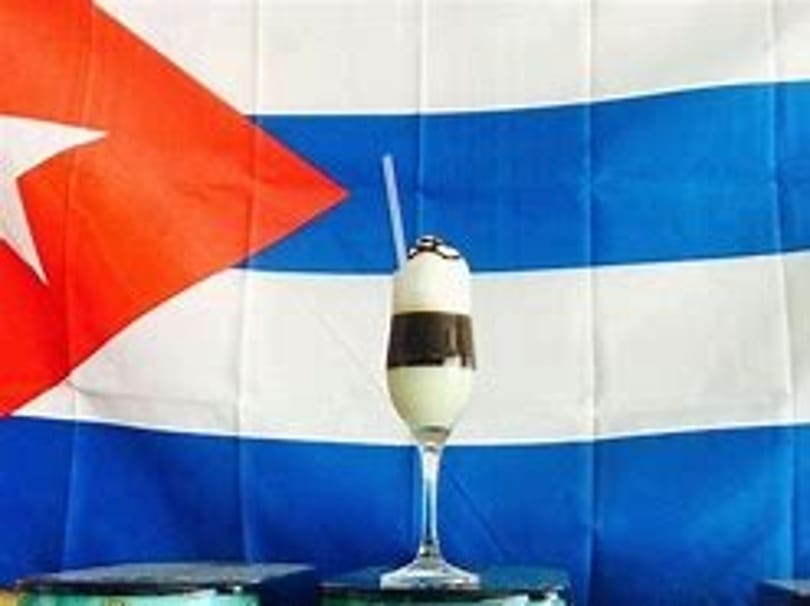 Layered white and brown drink with a straw in a tall glass, against a Cuban flag.