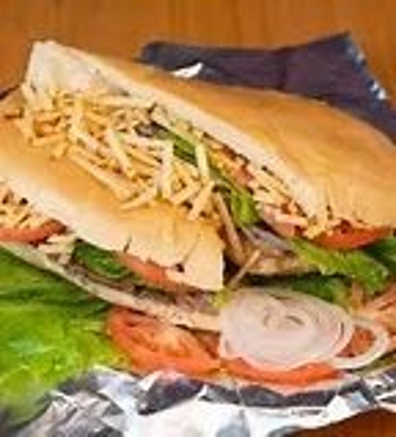 A large sandwich with lettuce, tomatoes, onions, and shoestring fries on foil.